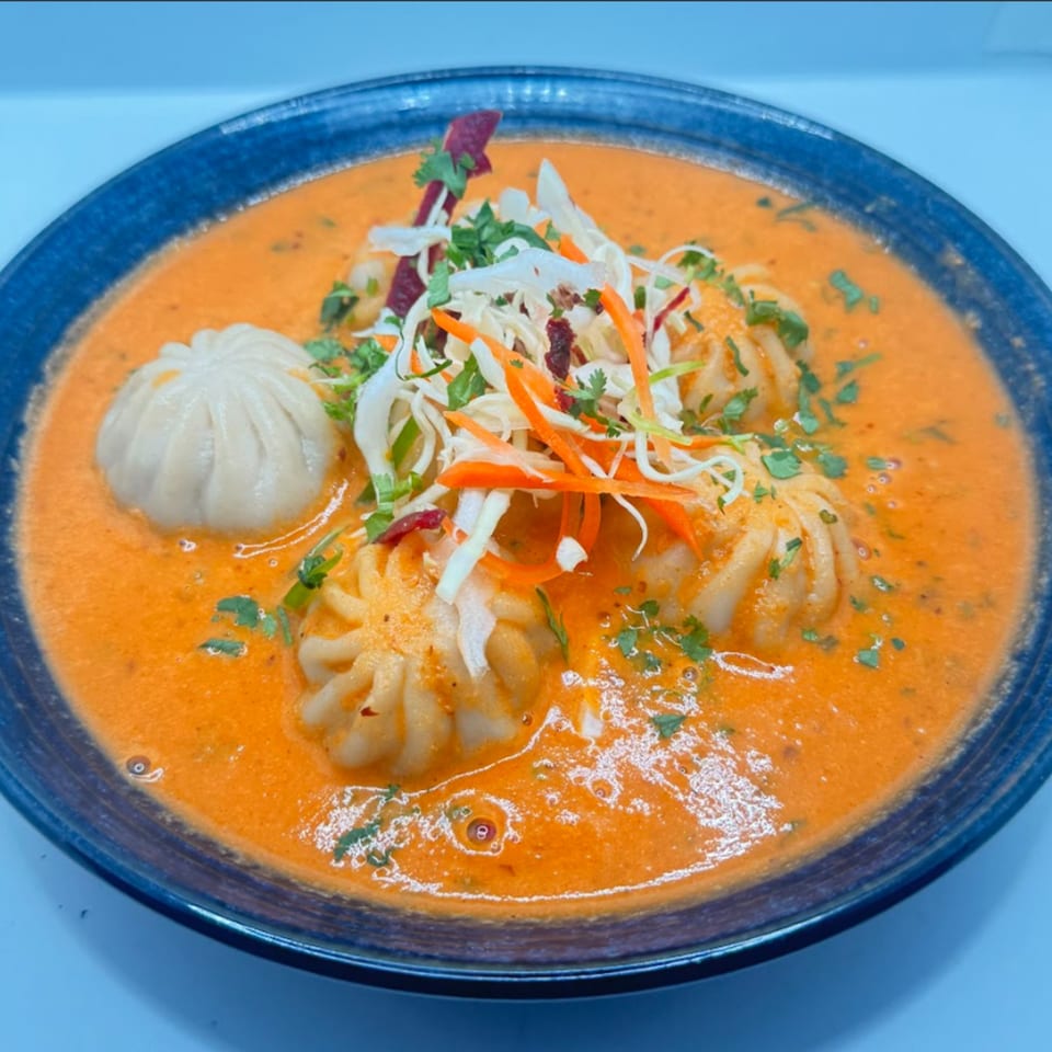 Delhi to Kathmandu's momo dumplings in a rich orange sauce, garnished with shredded vegetables and herbs.