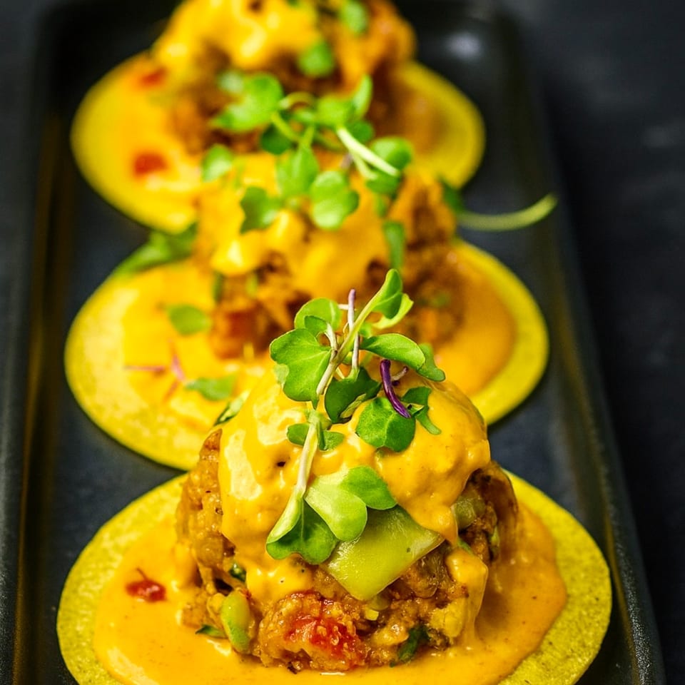 Three Indian tacos with yellow shells, orange sauce, and green garnish on a dark tray.